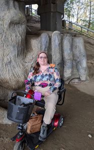 Naomi seated on a mobility scooter with red and purple trim. The Fremont Troll is behind her, with his hand over a VW bug.