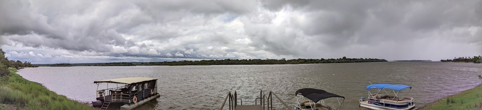 A panoramic view of the Zambezi river, with three boats and a warf in the foreground, and Zambia on the far shore.