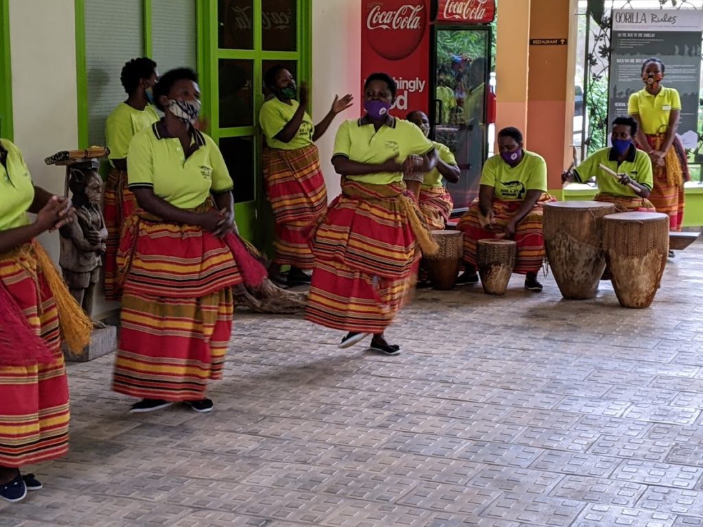 The dancers are on the left, and three drummers further back on the right. Shallon is standing behind the drummers.  All are wearing light green shirts and skirts with red and orange horizontal stripes