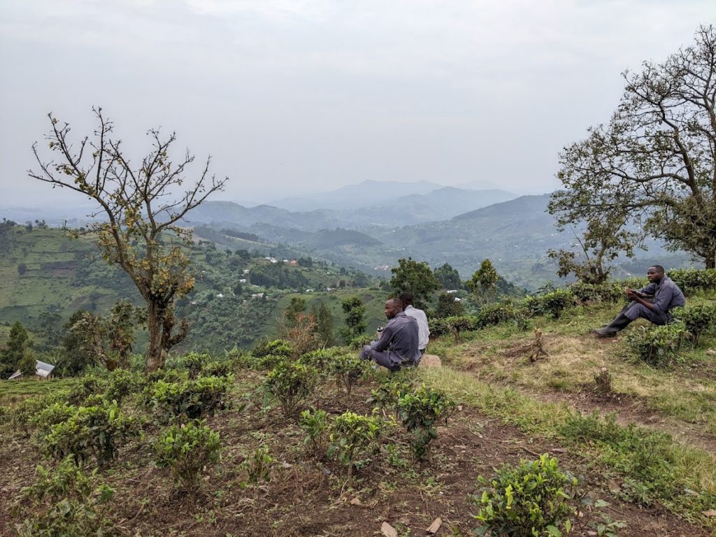 Three of my porters are sitting on the edge of a steep drop-off; there is a scattering of buildings in the middle distance.  In the background a series of mountains shrouded in mist.