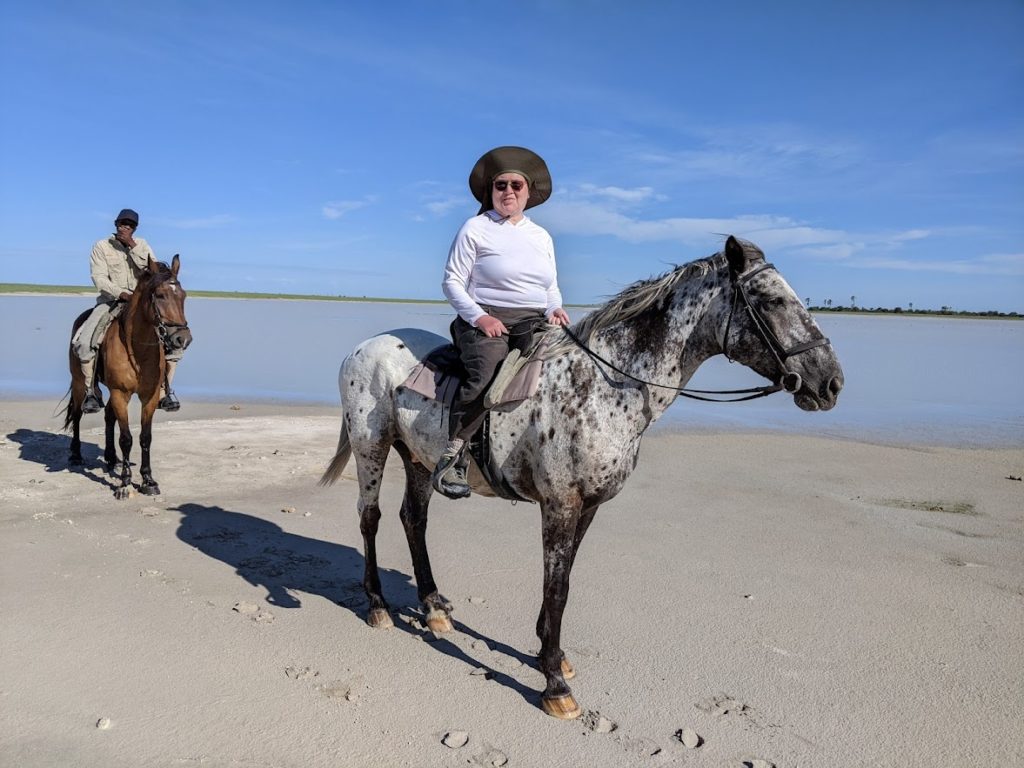 Naomi riding an Appaloosa horse -- white with black spots -- looking toward the camera. One of the guides is behind her riding a bay.