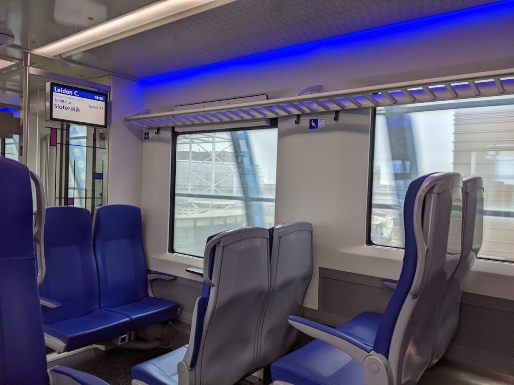 The inside of a train; there are three pairs of blue-upholstered seats on the far side, the farthest pair facing back toward the camera and the other two pairs facing forward.  The walls and backs of the seats are various shades of grey,  There is a luggage rack above the seats, and a strip of blue soffit lighting below the ceiling.