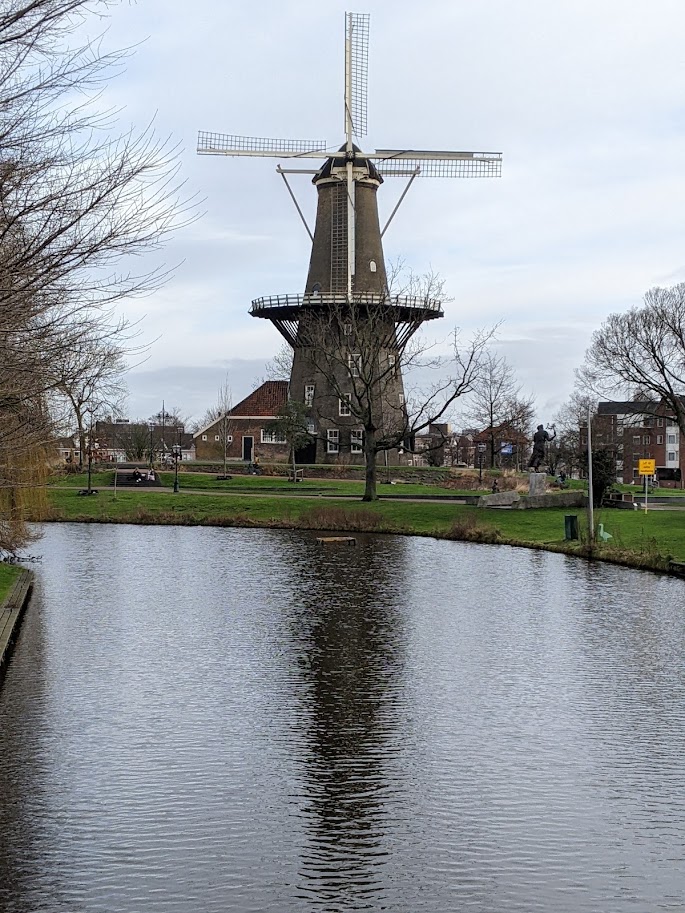 A windmill, with buildings behind it.  A canal occupies the entire bottom of the frame; its farther bank curves to the left.