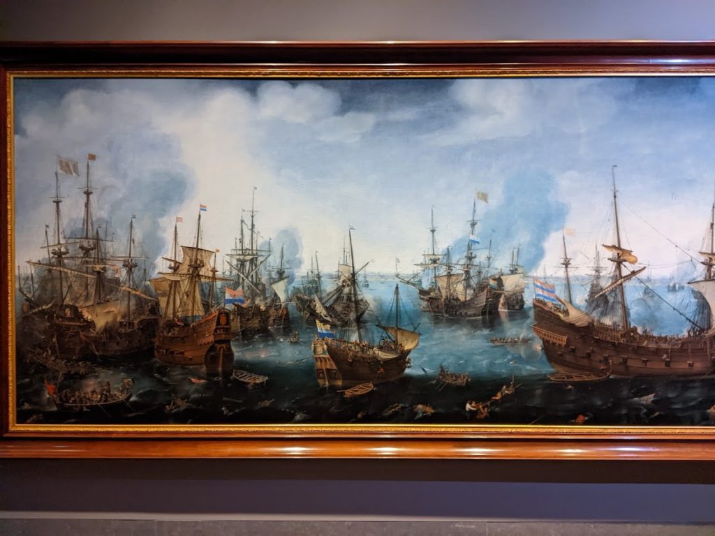 A wide painting in a wooden frame.  Most of the ships depicted are square-rigged.