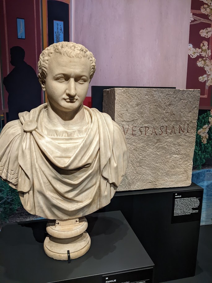 On the left, a bust of a man with short curly hair and a disapproving expression, on the left.  On the right, a block of stone on which is carved the name "Vespasiani".