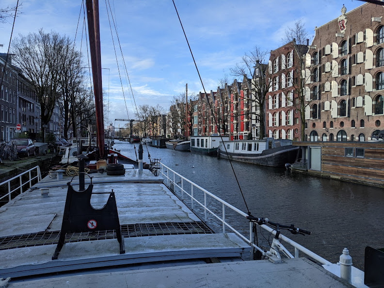 In the left foreground is the bow portion of my houseboat, with a bicycle parked by the starboard rail.  The canal is to the right, with a good view of houses and houseboats.