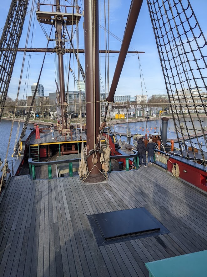 The Amsterdam's deck is made of grey planks; there is a railing with bright green pillars behind a brown mast; much of the other trim is brick-red.