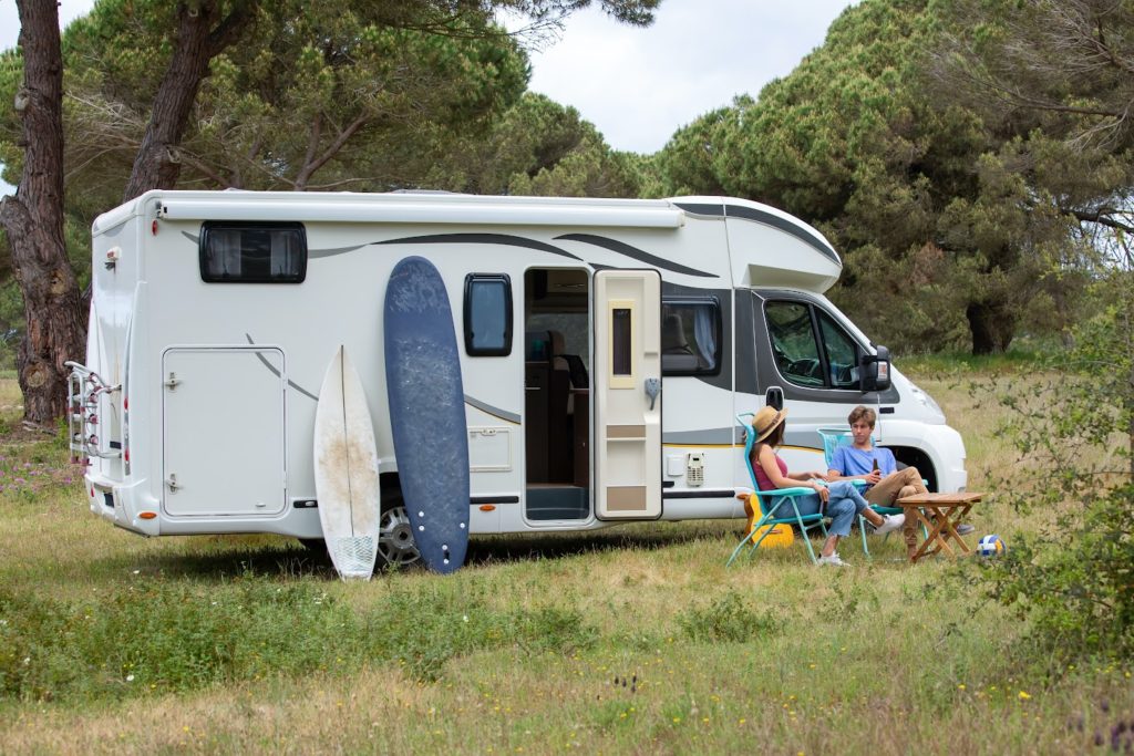 Stock photo of a Class C motor home; basically a van with an extended rear end and a section extending over the cab.  White with grey swooshes; there are two people in lawn chairs and two surfboards or paddleboards leaning against the side.