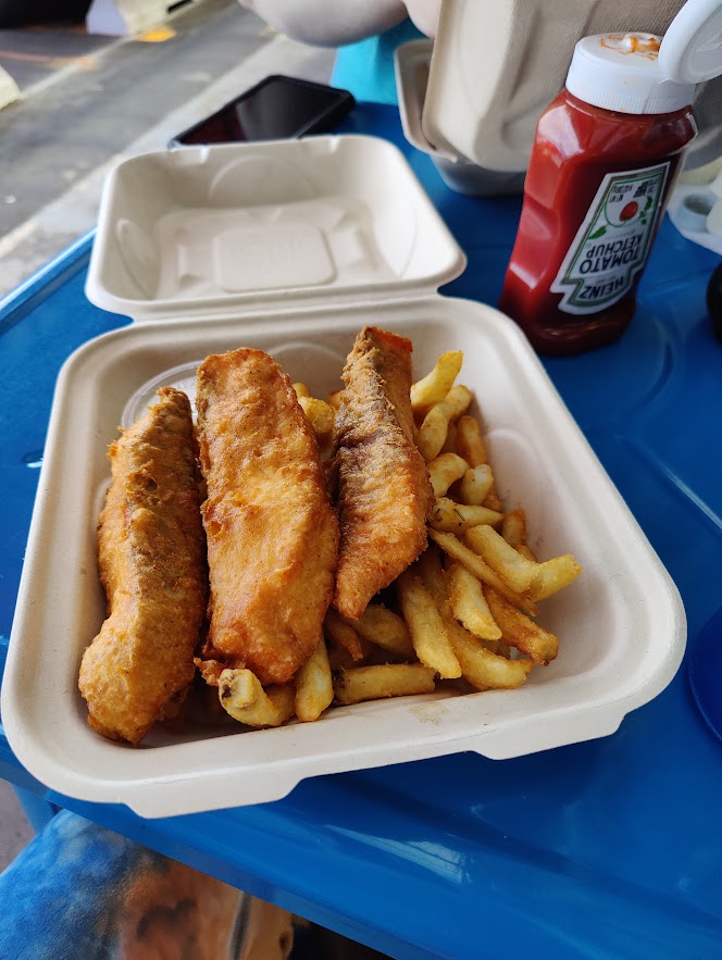Three pieces of fried fish on a bed of French fries, in a white clamshell take-out container.
