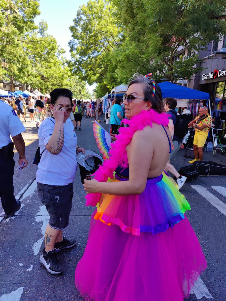 A woman in a pink backless dress with a rainbow overskirt, wearing a pink feather boa.
