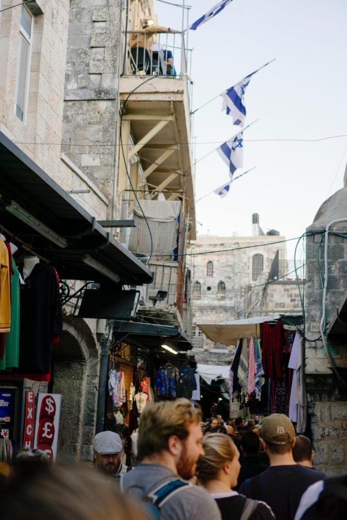 A narrow market street crowded with people.  Israeli flags fly overhead.
