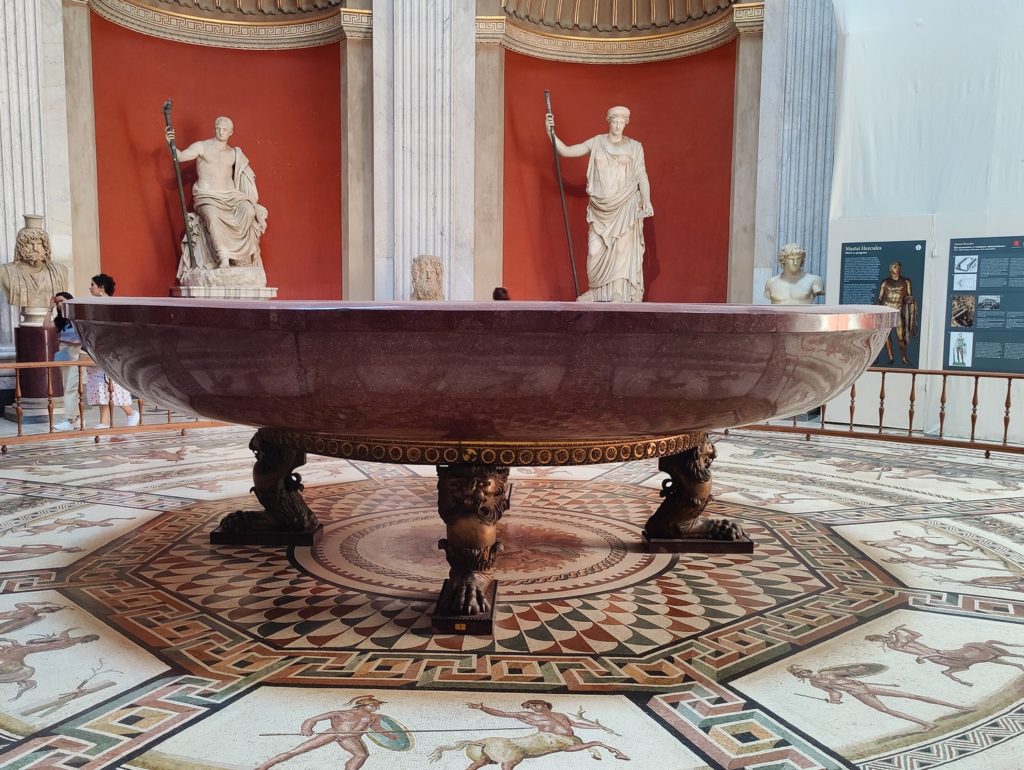 A huge bowl of reddish stone, standing on three legs on a tiled hexagon.  Two marble statues occupy niches in the background.