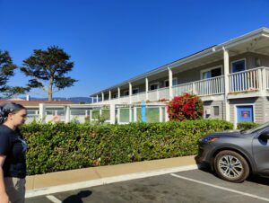 A two-story motel. C stands on the far left, in front of a hedge.