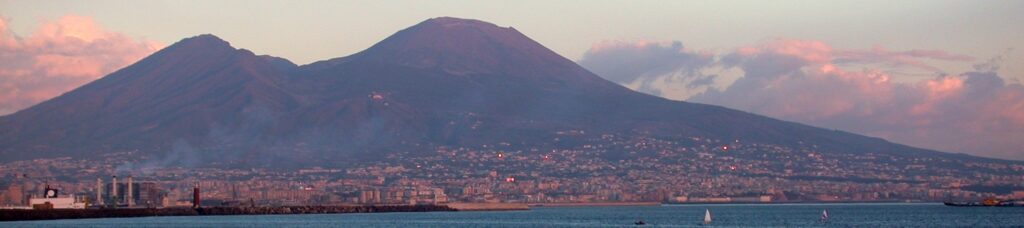 A volcano, with two peaks, at sunset.  A city sprawls dangerously close at its feet.